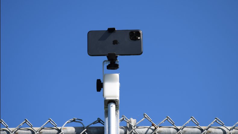 The QM-1 mount clamped to the top of a court fence, holding a phone above the fence line